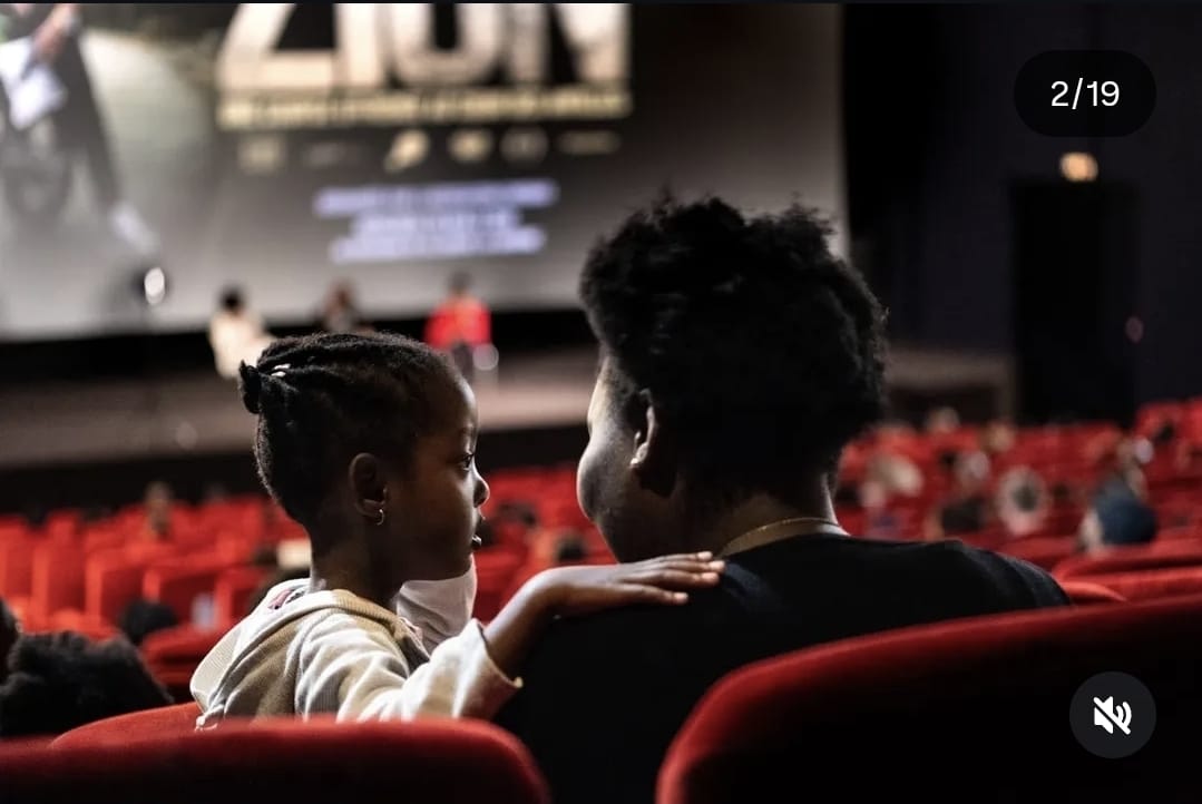 Une adulte et un enfant s'asseyent dans une salle de cinéma, regardant l'écran. L'enfant est habillé d'un pull clair et l'adulte de noir, avec un collier visible.